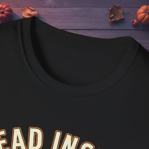 Close-up of a black crewneck t-shirt with a graphic reading 'HEAD IN THE COLD' in white and orange, laid flat on a dark purple wooden surface with autumn leaves and pumpkins.