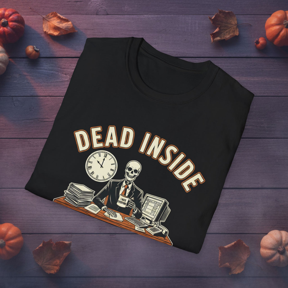 A black folded graphic T-shirt displayed on a dark purple wooden surface with autumn props like pumpkins and leaves. The shirt features a vintage-style illustration of a skeleton in a suit sitting at a desk with a computer, coffee mug, clock, and stacks of papers, with the words 'DEAD INSIDE' arched above.