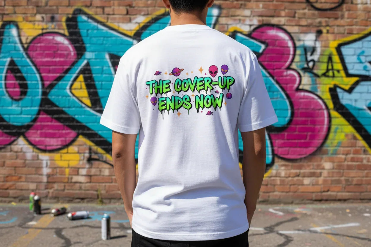 Man wearing white graphic t-shirt with 'The Cover-Up Ends Now' text and alien art, graffiti wall