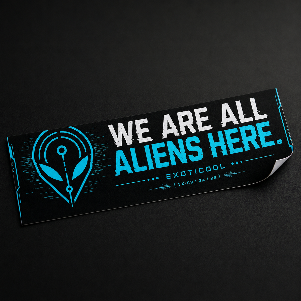 Angled view of black Exoticool “We Are All Aliens Here” bumper sticker with cyan alien artwork and a lifted corner.