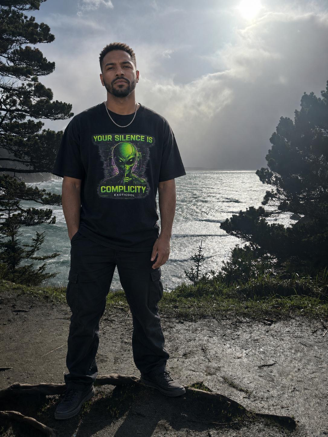 Adult model wearing the black Your Silence Is Complicity alien graphic tee at Cape Arago on the Oregon coast