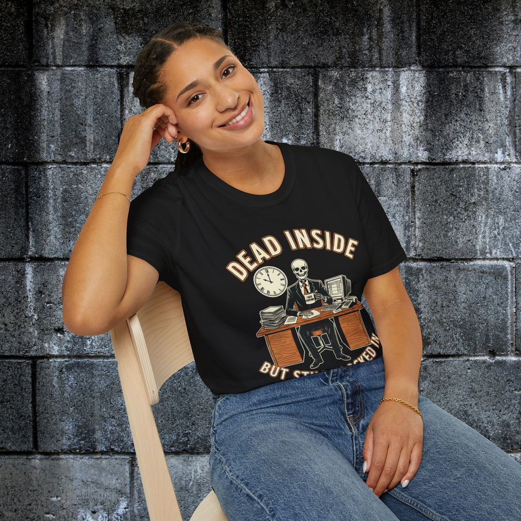 A woman sitting on a wooden chair against a dark gray brick wall, wearing a black graphic t-shirt with a skeleton office worker design and the text 'DEAD INSIDE BUT STAY clocked in'.