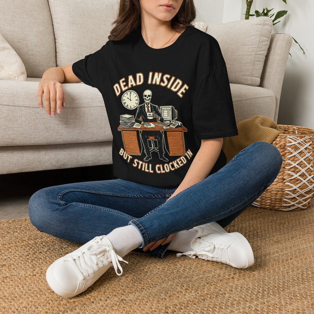 A woman sitting cross-legged on a woven rug in front of a beige sofa, wearing a black graphic T-shirt with a skeleton clock graphic and the text 'DEAD INSIDE BUT STILL CLOCKED IN'.