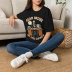 A woman sitting cross-legged on a woven rug in front of a beige sofa, wearing a black graphic T-shirt with a skeleton clock graphic and the text 'DEAD INSIDE BUT STILL CLOCKED IN'.