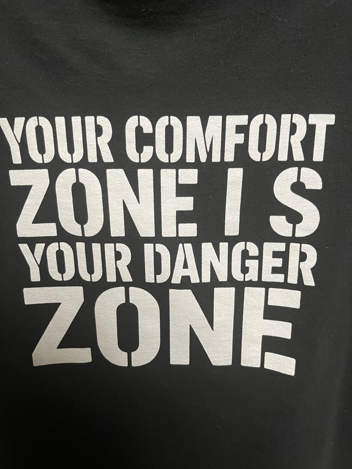 Close-up of Your Comfort Zone Is Your Danger Zone text on the back of the black tee