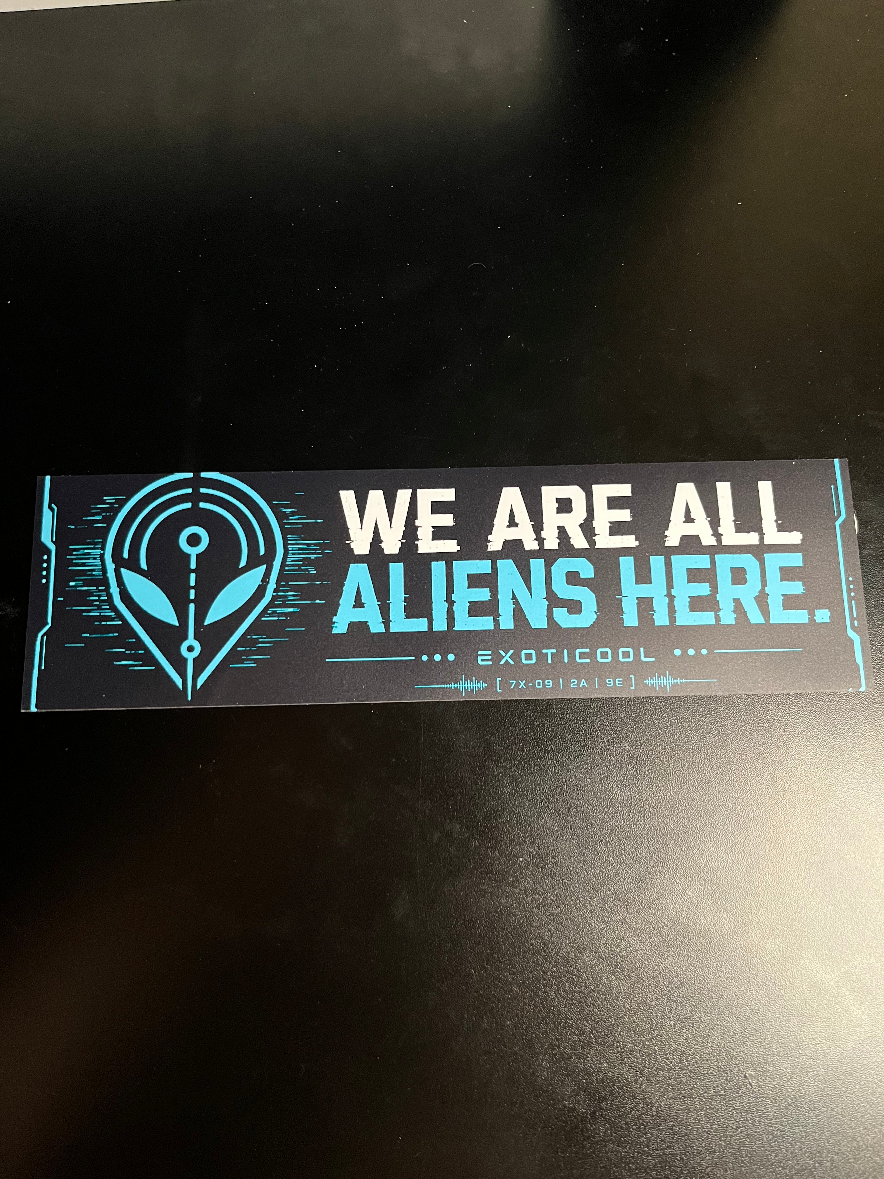 Real 11-inch We Are All Aliens Here bumper sticker displayed flat on a black surface