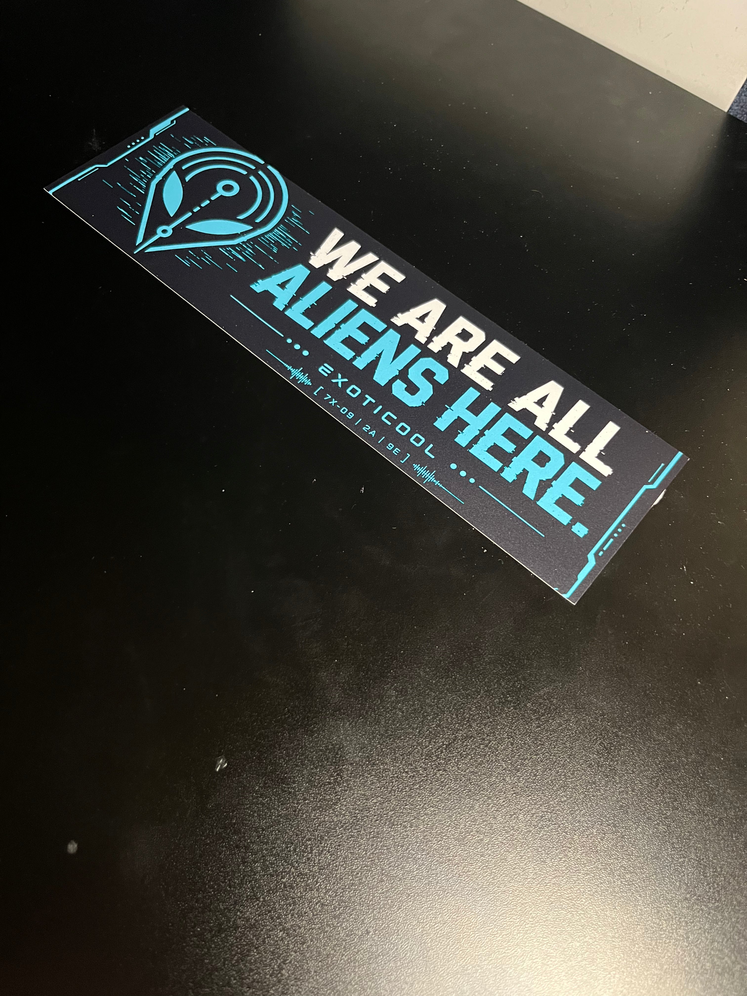 Angled view of the We Are All Aliens Here alien bumper sticker on a black tabletop