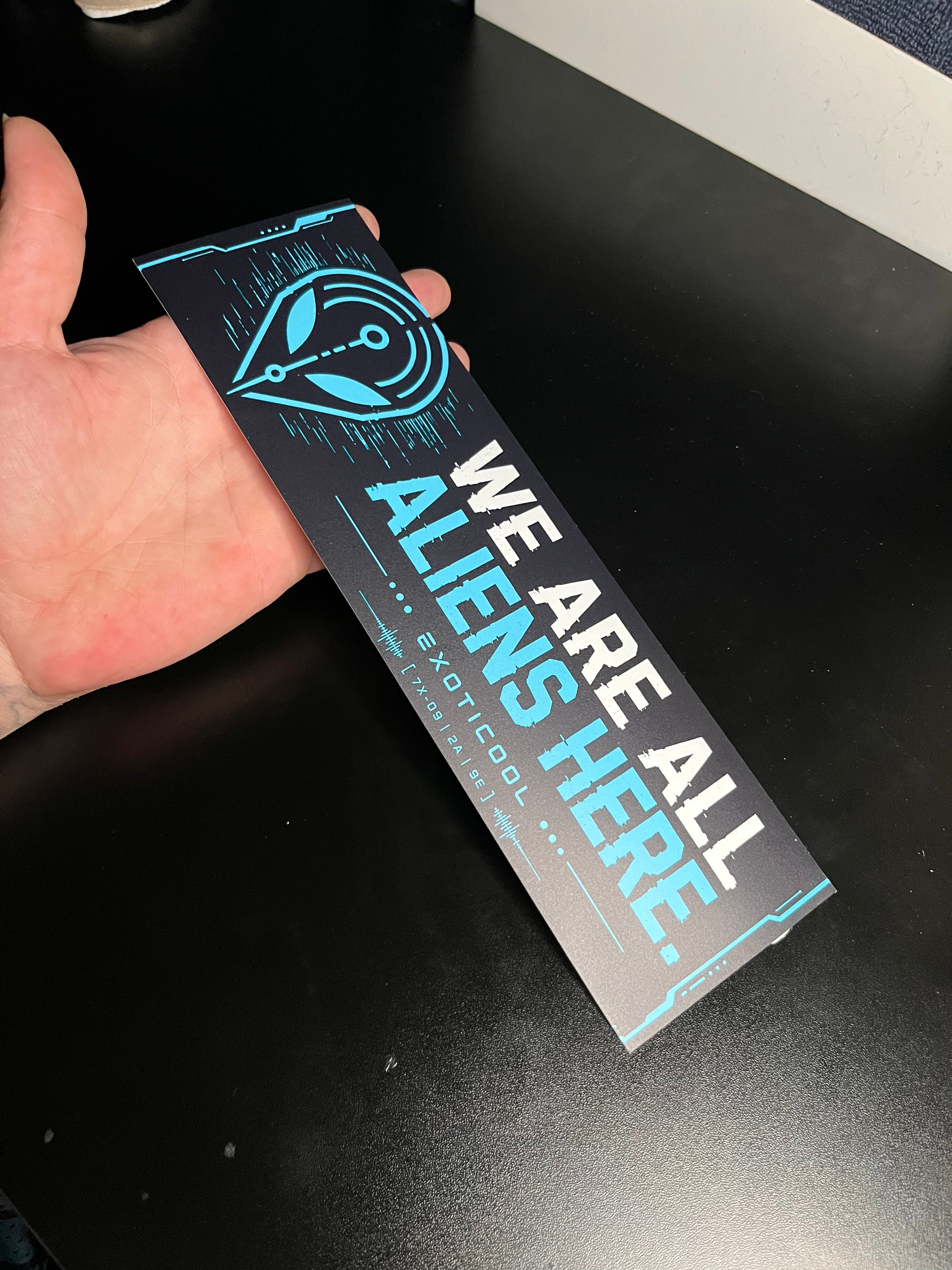 Hand holding the 11-inch We Are All Aliens Here bumper sticker to demonstrate its size