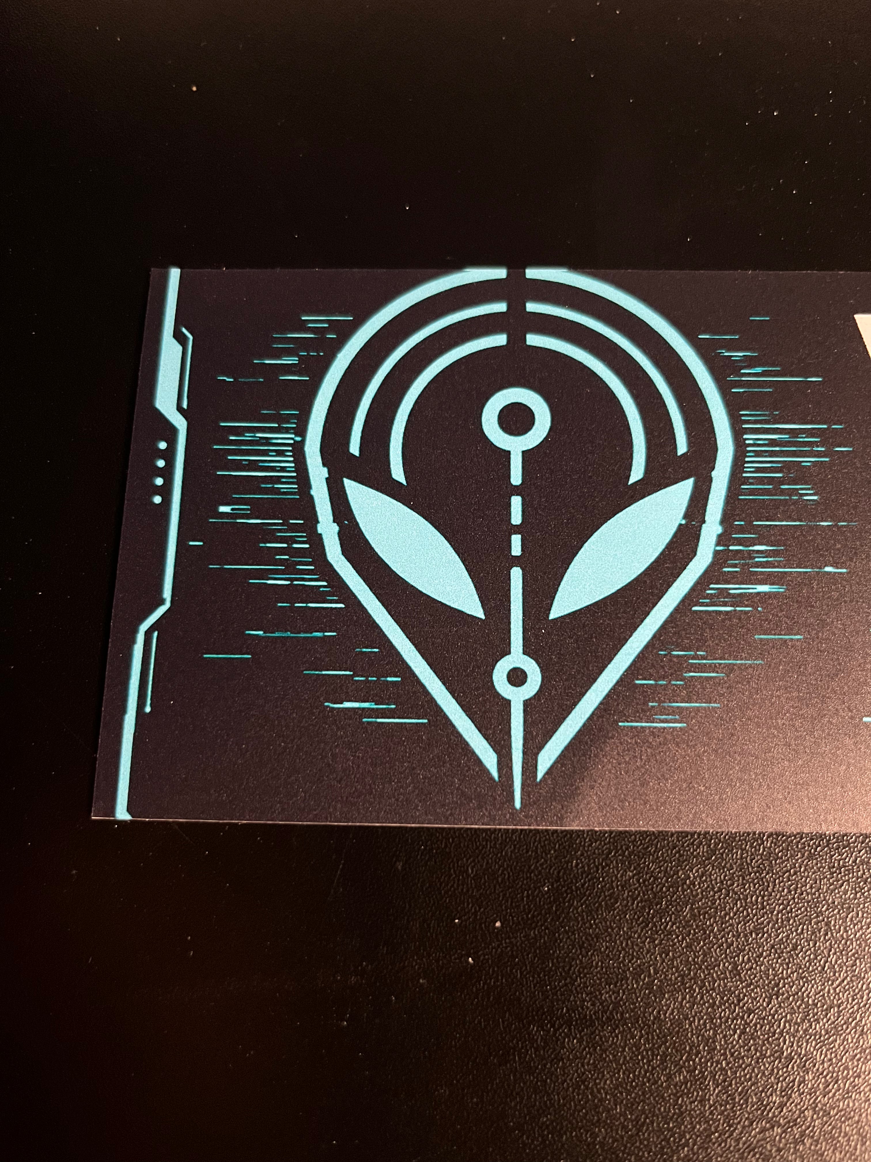 Close-up of the cyan alien signal emblem printed on the black bumper sticker