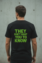 Back view of male model wearing black Exoticool Alien Signal Tee with large green stencil “They Don’t Want You To Know” lettering in an urban setting