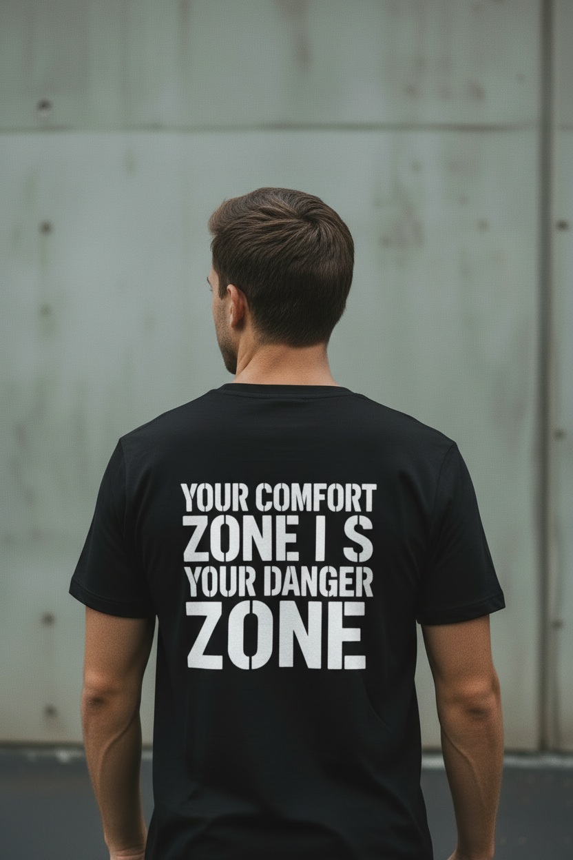 Back view of male model wearing black Exoticool System Glitch Tee with large white “Your Comfort Zone Is Your Danger Zone” lettering in an urban setting