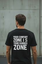Back view of male model wearing black Exoticool System Glitch Tee with large white “Your Comfort Zone Is Your Danger Zone” lettering in an urban setting